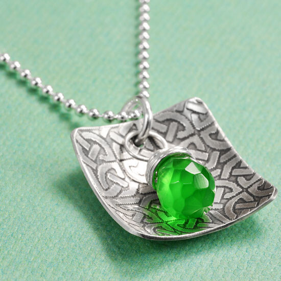 I love the textured pattern on the silver Close up view of a silver chain with a silver charm textured in a Celtic knot pattern, with a green faceted briolette